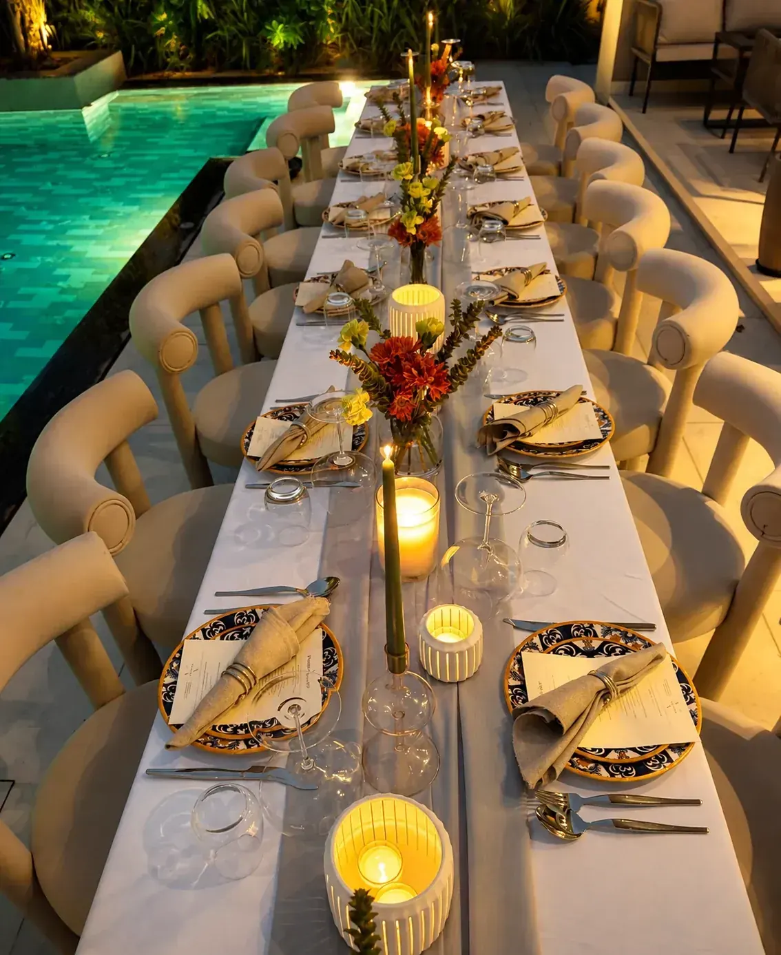 A long poolside table set for dinner at dusk with candlelight, printed menus and hand-painted plates