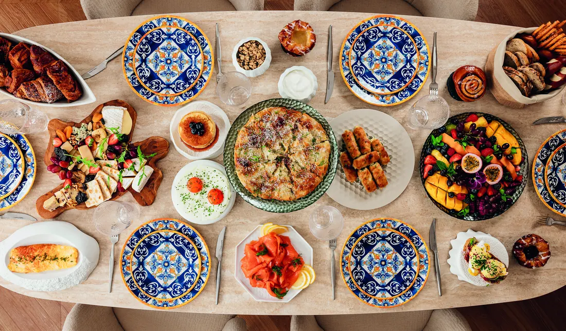 A dining table laid for six with hand-painted majolica plates, pastries, pancakes, cheeses and fruit, seen from above