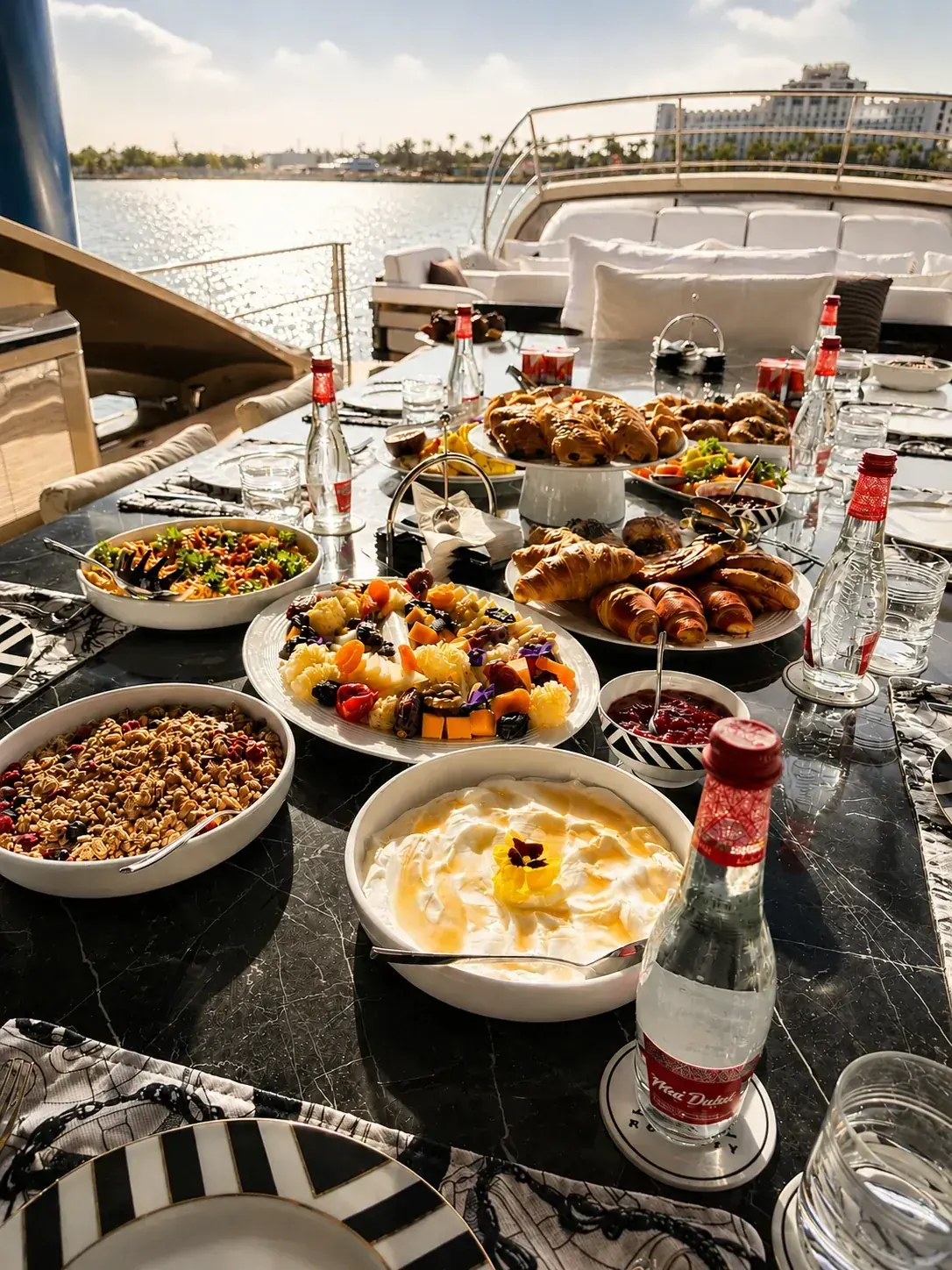A breakfast spread laid on a yacht deck table with pastries, fruit, granola and yoghurt, the marina behind